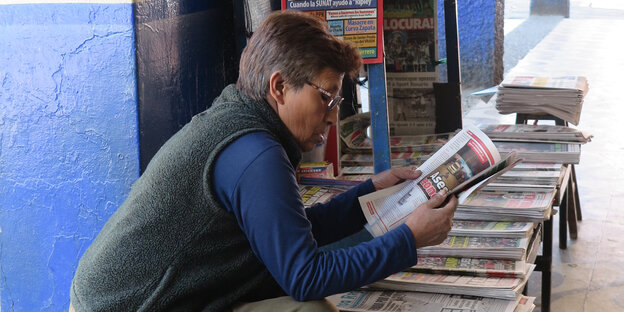 Frau sitzt über eine Zeitung gebeugt, lesend, dahinter ein Stand mit Zeitungen auf Spanisch Frau sitzt über eine Zeitung gebeugt, lesend, dahinter ein Stand mit Zeitungen auf Spanisch