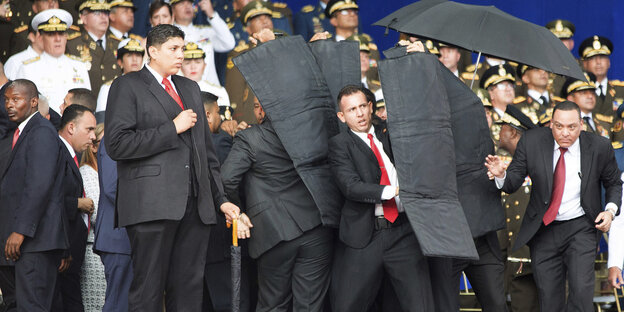 Menschen in Uniformen, in der Mitte wird Nicolas Maduro von Soldaten mit kugelsicheren Planen geschützt Menschen in Uniformen, in der Mitte wird Nicolas Maduro von Soldaten mit kugelsicheren Planen geschützt