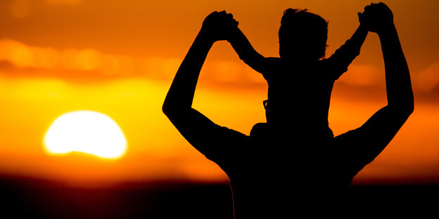 Ein Vater steht mit seinem Sohn auf den Schultern vor der untergehenden Sonne