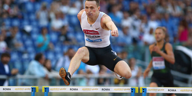 Karsten Warholm springt in Sportbekleidung über eine Hürde, dahinter sitzen Zuschauer in einem Stadion Karsten Warholm springt in Sportbekleidung über eine Hürde, dahinter sitzen Zuschauer in einem Stadion