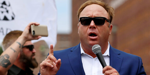 Alex Jones trägt einen Anzug und eine Sonnenbrille und spricht mit einem Mikrofon in der Hand, im Hintergrund schießt ein bärtiger Demonstrant ein Foto