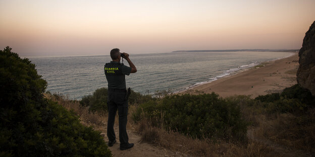 Ein Mann steht an einer Küste und guckt durch ein Fernglas auf das Meer Ein Mann steht an einer Küste und guckt durch ein Fernglas auf das Meer