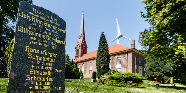 , im Vordergrund ein Familiengrabstein. Hinter dem Gebäude ragt eine Windkraftanlage hervor.