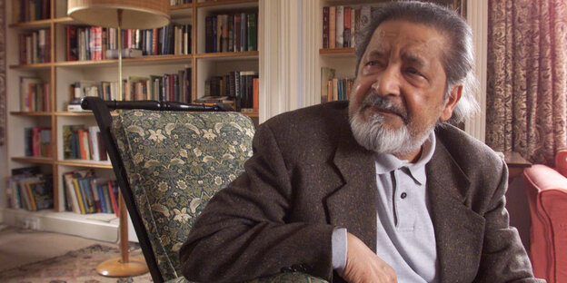 Ein älterer Mann sitzt in einem Stuhl, im Hintergrund viele Bücher in Regalen. Es ist der Literaturnobelpreisträger Naipaul