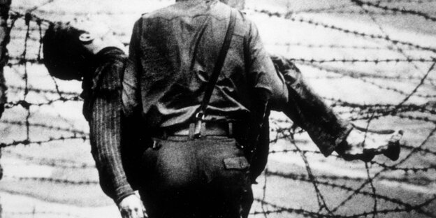 Schwarz-weiß-Foto: Ein Uniformierter trägt einen Mann vor Stacheldraht Schwarz-weiß-Foto: Ein Uniformierter trägt einen Mann vor Stacheldraht
