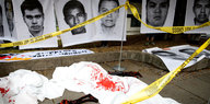 Waffenattrapen und rote Flecken auf dem Boden, dahinter Bilder junger Mexikaner