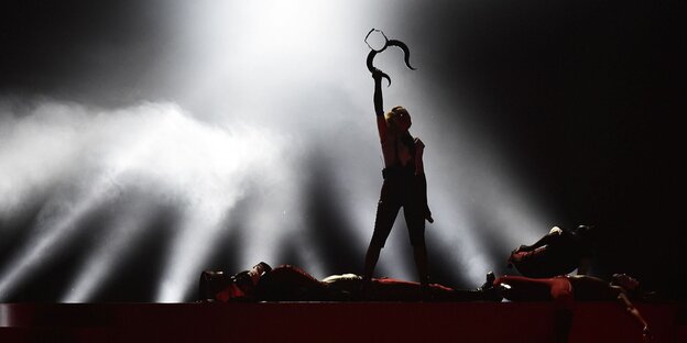 Die Silhouette von Madonna steht auf einer Bühne und hält einen gehörnten Kopfaufsatz in die Höhe, sie wird von unten angestrahlt Die Silhouette von Madonna steht auf einer Bühne und hält einen gehörnten Kopfaufsatz in die Höhe, sie wird von unten angestrahlt