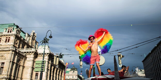 Ein Mann in Badehose steht auf einem Truck, er trägt einen pinken Hut und eine Blumenkette, an seinem Rücken prangen regenbogenfarbene Flügel Ein Mann in Badehose steht auf einem Truck, er trägt einen pinken Hut und eine Blumenkette, an seinem Rücken prangen regenbogenfarbene Flügel