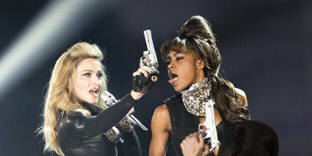 Madonna steht auf einer Bühne, hält einen silbernen Revolver in der Hand, neben ihr eine Tänzerin Madonna steht auf einer Bühne, hält einen silbernen Revolver in der Hand, neben ihr eine Tänzerin