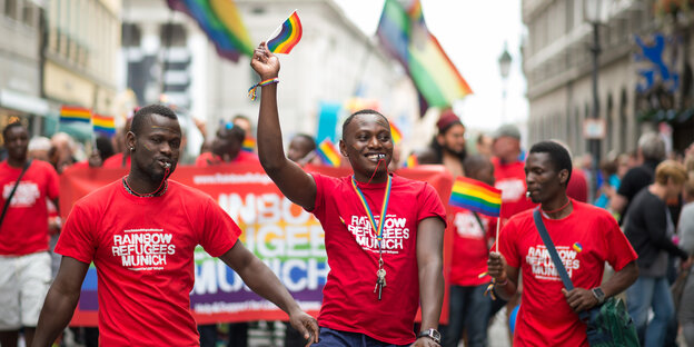 Drei Männer mit T-Shirts der „Rainbow Refugees Munich“, einer hält eine Regenbogenfahne Drei Männer mit T-Shirts der „Rainbow Refugees Munich“, einer hält eine Regenbogenfahne
