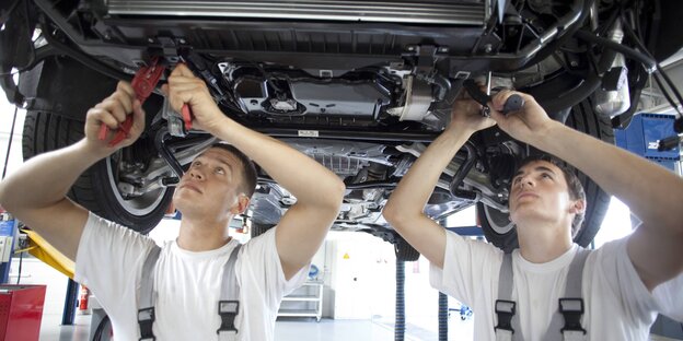 Zwei Männer in Arbeitskleidung stehen mit Werkzeug in der Hand unter einem Auto Zwei Männer in Arbeitskleidung stehen mit Werkzeug in der Hand unter einem Auto
