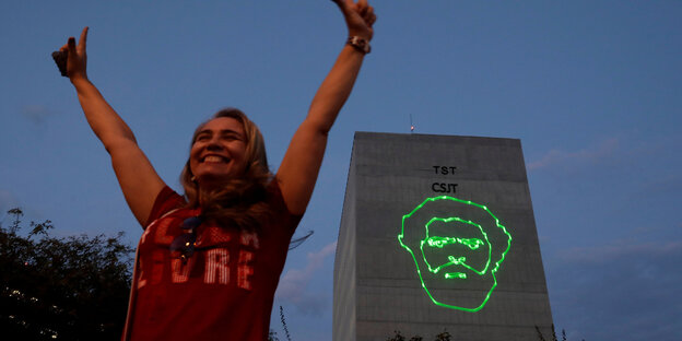 Eine Frau streckt erfreut die Arme in die Höhe, im Hintergrund ist das Gesicht von Lula mit grünen Lasern an eine Hauswand projeziert Eine Frau streckt erfreut die Arme in die Höhe, im Hintergrund ist das Gesicht von Lula mit grünen Lasern an eine Hauswand projeziert