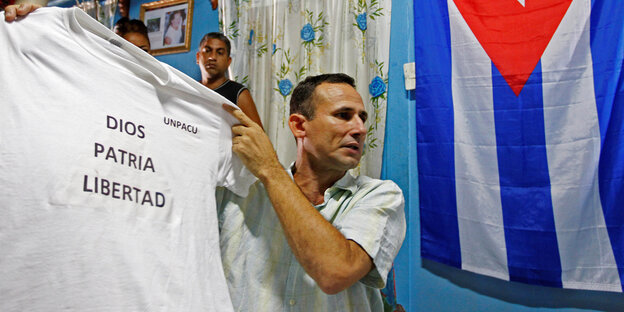 Der kubanische Dissident José Daniel Ferrer hält vor einer kubanischen Flagge ein T-Shirt mit der Aufschrift „Gott, Heimat, Freiheit“ in die Höhe Der kubanische Dissident José Daniel Ferrer hält vor einer kubanischen Flagge ein T-Shirt mit der Aufschrift „Gott, Heimat, Freiheit“ in die Höhe