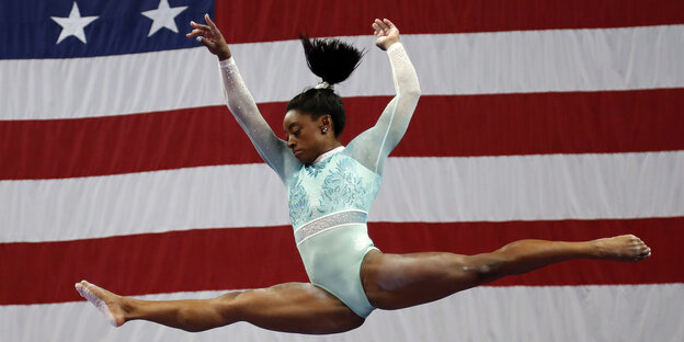 Simone Biles in einem Spagat-Sprung vor US-Flagge