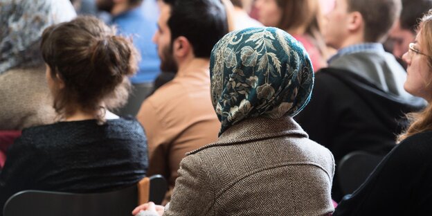 Eine Frau mit Kopftuch sitzt zwischen anderen Männern und Frauen, sie alle sitzen auf Stühlen, sind von hinten zu sehen und schauen geradeaus. Eine Frau mit Kopftuch sitzt zwischen anderen Männern und Frauen, sie alle sitzen auf Stühlen, sind von hinten zu sehen und schauen geradeaus.