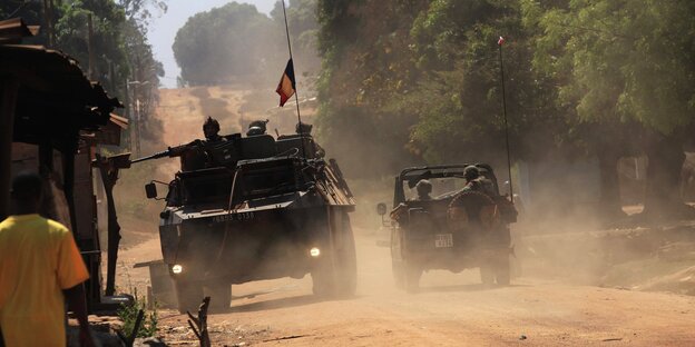 Ein Radpanzer mit französischer Flagge auf staubiger Straße in Bangui. Soldaten mit Schusswaffen darauf. Daneben ein Jeep. Links auf der Straße läuft ein Mann in gelbem Shirt. Ein Radpanzer mit französischer Flagge auf staubiger Straße in Bangui. Soldaten mit Schusswaffen darauf. Daneben ein Jeep. Links auf der Straße läuft ein Mann in gelbem Shirt.