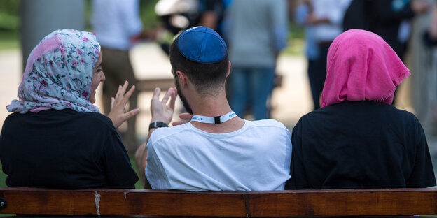 Zwei junge Muslimas mit Kopftuch und ein junger jüdischer Mann mit Kippa unterhalten sich miteinander Zwei junge Muslimas mit Kopftuch und ein junger jüdischer Mann mit Kippa unterhalten sich miteinander