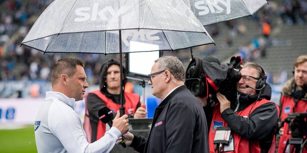 Zwei Männer auf einem Fußballplatz unter einem durchsichtigen Regenschirm. Der eine hält dem anderen ein Mikrofon vor das Gesicht Zwei Männer auf einem Fußballplatz unter einem durchsichtigen Regenschirm. Der eine hält dem anderen ein Mikrofon vor das Gesicht