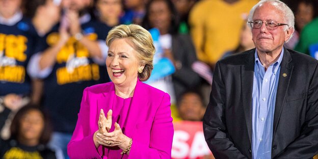 Hillary Clinton vor Publikum - lachend und in die Hände klatschend. Dahinter versetzt steht mit ernster Miene Bernie Sanders Hillary Clinton vor Publikum - lachend und in die Hände klatschend. Dahinter versetzt steht mit ernster Miene Bernie Sanders