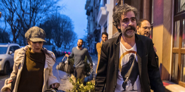 Yücel und seine Frau laufen über eine Straße, Männer im Hintergrund Yücel und seine Frau laufen über eine Straße, Männer im Hintergrund