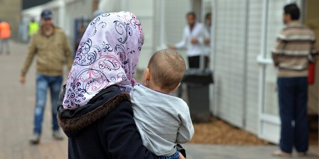 Eine Kopftuch tragende Frau hält ein Kind auf dem Arm; im Hintergrund Männer Eine Kopftuch tragende Frau hält ein Kind auf dem Arm; im Hintergrund Männer