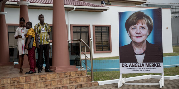 Ein PLakat, das Angela Merkel zeigt, steht neben einem Flughafengebäude. Neben dem Plakat stehen drei Menschen Ein PLakat, das Angela Merkel zeigt, steht neben einem Flughafengebäude. Neben dem Plakat stehen drei Menschen