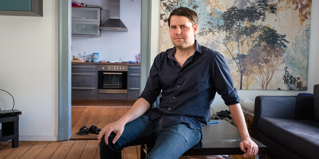 Ein Mann in Jeans und dunklem Hemd sitzt auf dem Couchtisch in einer Wohngung Ein Mann in Jeans und dunklem Hemd sitzt auf dem Couchtisch in einer Wohngung