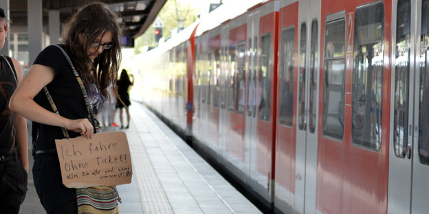 Eine Frau steht auf einem Bahnsteig. In ihrer Hand hält sie ein Schild, auf dem steht, dass sie schwarz fährt Eine Frau steht auf einem Bahnsteig. In ihrer Hand hält sie ein Schild, auf dem steht, dass sie schwarz fährt