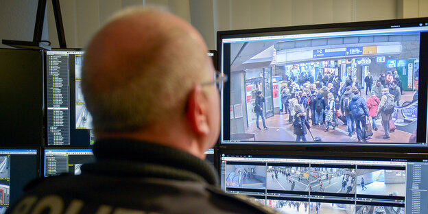 Ein Polizist betrachtet einen Monitor auf dem eine Menschenmenge zu sehen ist. Ein Polizist betrachtet einen Monitor auf dem eine Menschenmenge zu sehen ist.