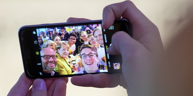 Hände halten ein Handy - darauf ein Foto mit Angela Merkel und Peter Tauber, umringt von Kindern Hände halten ein Handy - darauf ein Foto mit Angela Merkel und Peter Tauber, umringt von Kindern