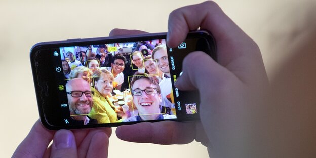 Hände halten ein Handy - darauf ein Foto mit Angela Merkel und Peter Tauber, umringt von Kindern