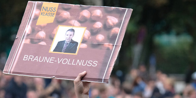 Aus einer Menschenmenge wird ein Plakat im Stil einer Schokoladenpackung und mit der Aufschrift „Braune Vollnuss“ gezeigt Aus einer Menschenmenge wird ein Plakat im Stil einer Schokoladenpackung und mit der Aufschrift „Braune Vollnuss“ gezeigt