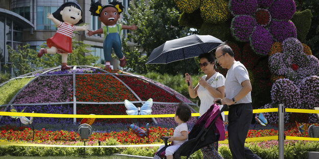 Vor einer Installation mit bunten Blumen sowie einer asiatischen und einer schwarzen Mädchenfigur läuft eine Familie mit Kinderwagen Vor einer Installation mit bunten Blumen sowie einer asiatischen und einer schwarzen Mädchenfigur läuft eine Familie mit Kinderwagen