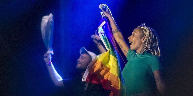 Drei Menschen winken, einer hat eine Regenbogenfahne in der Hand