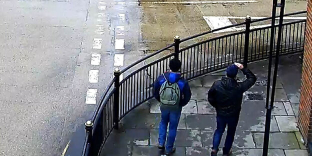 An einer Straße stehen zwei Männer mit Mützen An einer Straße stehen zwei Männer mit Mützen