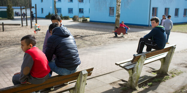 Kinder sitzen gelangweilt auf Bänken an einem Spielplatz vor kasernenartigen Gebäuden Kinder sitzen gelangweilt auf Bänken an einem Spielplatz vor kasernenartigen Gebäuden