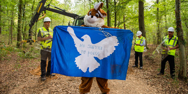 Ein Aktivist im Hasenkostüm und mit Friedenstaubenflagge in den Händen steht vor einem Bagger und RWE-Mitarbeitern