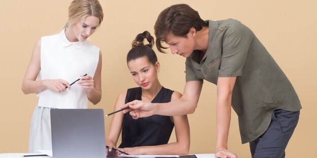 Eine Frau sitzt vor einem Laptop, rechts und links neben ihr stehen eine Frau und ein Mann, der auf den Bildschirm zeigt Eine Frau sitzt vor einem Laptop, rechts und links neben ihr stehen eine Frau und ein Mann, der auf den Bildschirm zeigt