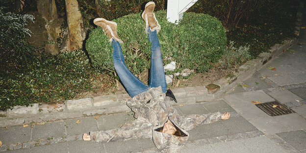 Gestelltes Foto einer Person, die auf dem Gehweg auf dem Rücken liegt, die Füße in einer Hecke Gestelltes Foto einer Person, die auf dem Gehweg auf dem Rücken liegt, die Füße in einer Hecke