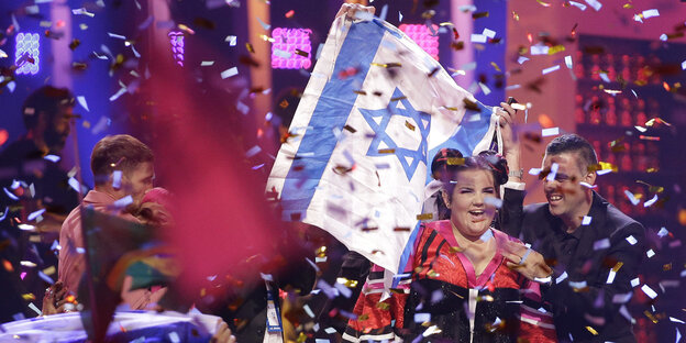 In einem Konfettiregen steht eine junge Frau, hinter ihr die israelische Flagge In einem Konfettiregen steht eine junge Frau, hinter ihr die israelische Flagge
