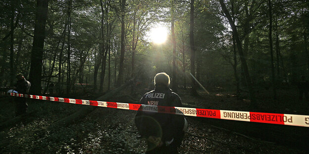 Ein Polizist steht vor einem rot-weißen Flatterband im Wald und guckt ins Sonnendurchflutete Dickicht Ein Polizist steht vor einem rot-weißen Flatterband im Wald und guckt ins Sonnendurchflutete Dickicht