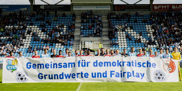 Fußballer stehen auf dem Rasen und halten ein Transparent mit der Aufschrift „Gemeinsam für demokratische Grundwerte und Fairplay“ Fußballer stehen auf dem Rasen und halten ein Transparent mit der Aufschrift „Gemeinsam für demokratische Grundwerte und Fairplay“