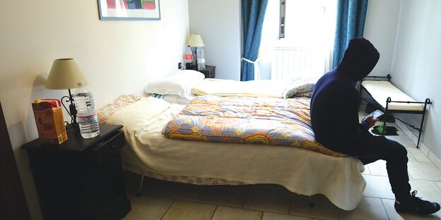 Ein junger Mensch mit Kapuzenpulli sitzt auf einem Bett. Ein junger Mensch mit Kapuzenpulli sitzt auf einem Bett.