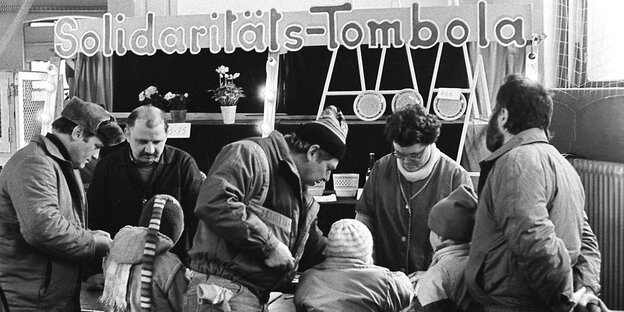Ein schwarz-weiß-Foto: Mehrere Männer stehen mit Kindern an einem Stand mit der Aufschrift "Solidaritäts-Tombola" Ein schwarz-weiß-Foto: Mehrere Männer stehen mit Kindern an einem Stand mit der Aufschrift "Solidaritäts-Tombola"