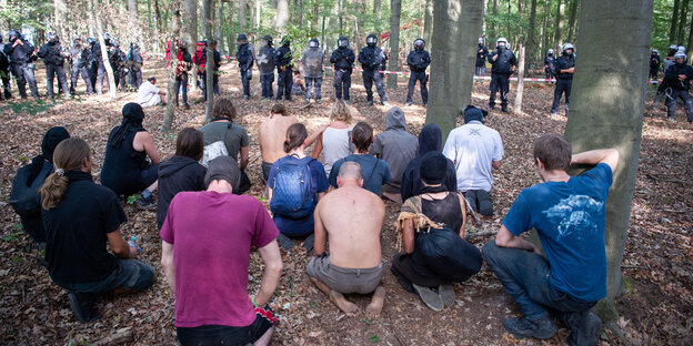 Menschen knien im Wald, davor bilden Polizisten in Uniform eine Kette Menschen knien im Wald, davor bilden Polizisten in Uniform eine Kette