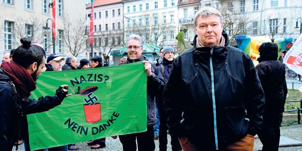 Kundgebung mit antifaschistischem Transparent, im Vordergund schaut ein Mann in die Kamera Kundgebung mit antifaschistischem Transparent, im Vordergund schaut ein Mann in die Kamera