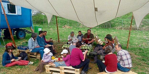 Eine Gruppe Menschen sitzt unter einem Sonnensegel auf Paletten und isst. Eine Gruppe Menschen sitzt unter einem Sonnensegel auf Paletten und isst.