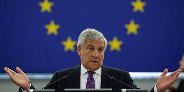 Tajani sitzt von einer EU-Flagge und macht eine fragende Geste Tajani sitzt von einer EU-Flagge und macht eine fragende Geste
