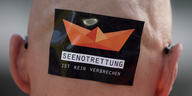 Ein Mann mit einem Aufkleber auf dem Kopf. Darauf steht: "Seenotrettung ist kein Verbrechen". Ein Mann mit einem Aufkleber auf dem Kopf. Darauf steht: "Seenotrettung ist kein Verbrechen".