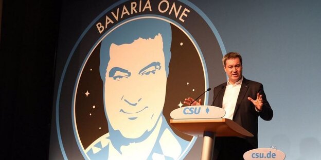 Markus Söder steht an einem Pult, dahinter ein Logo mit Söders Gesicht und Schriftzug "Bavaria One" Markus Söder steht an einem Pult, dahinter ein Logo mit Söders Gesicht und Schriftzug "Bavaria One"